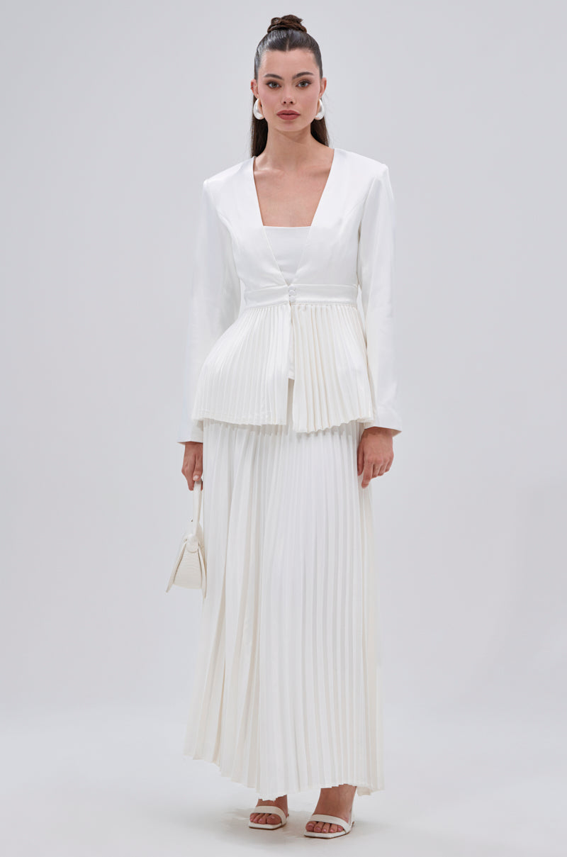 A woman stands against a plain background wearing a white long-sleeve blazer with a pleated peplum, the SWEET THING PLEATED SATIN MAXI SKIRT, white heeled sandals, and holding a small white handbag. 9