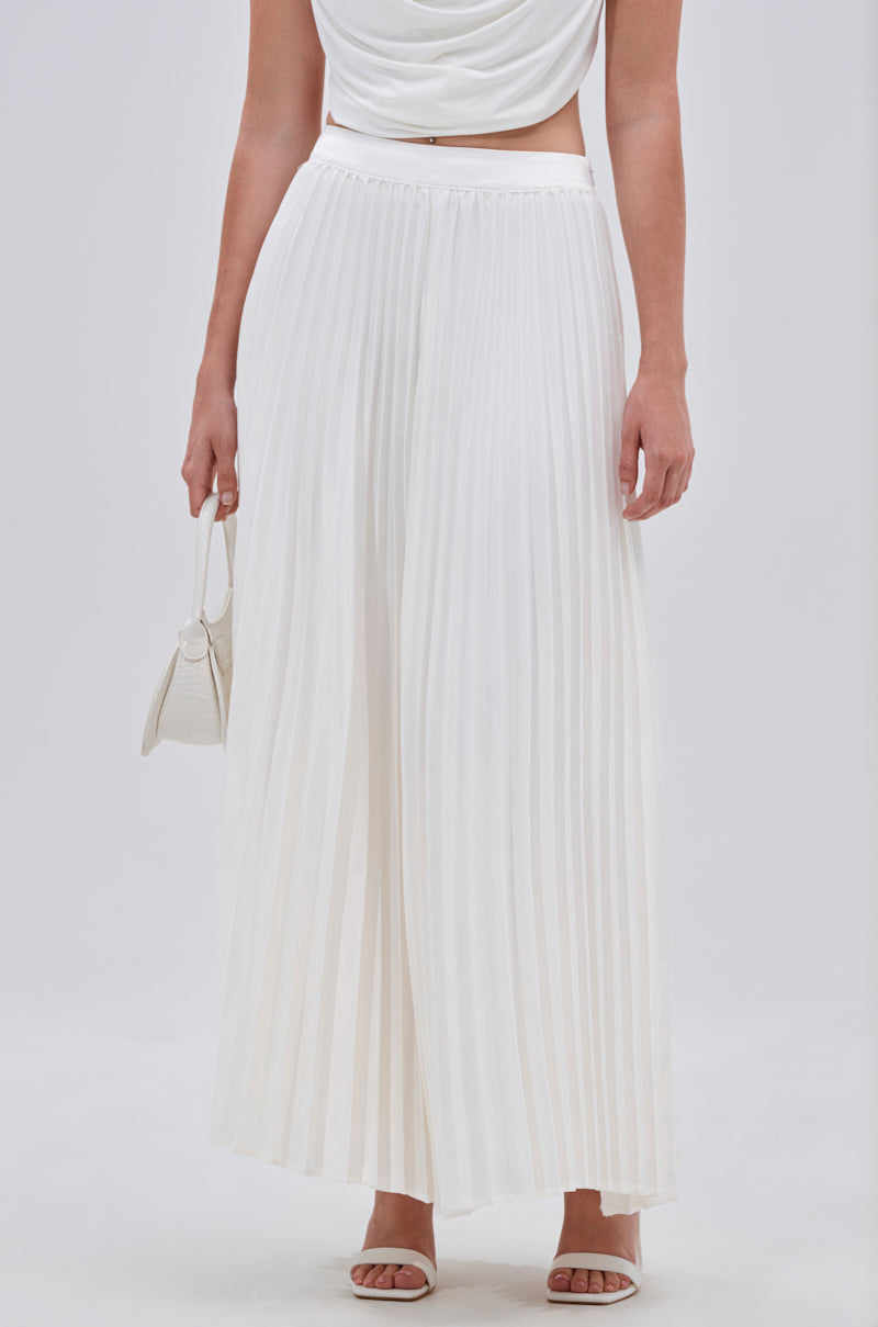 A woman wearing the SWEET THING PLEATED SATIN MAXI SKIRT, paired with a white sleeveless top, white heeled sandals, and a small white handbag, stands against a plain light background. 0