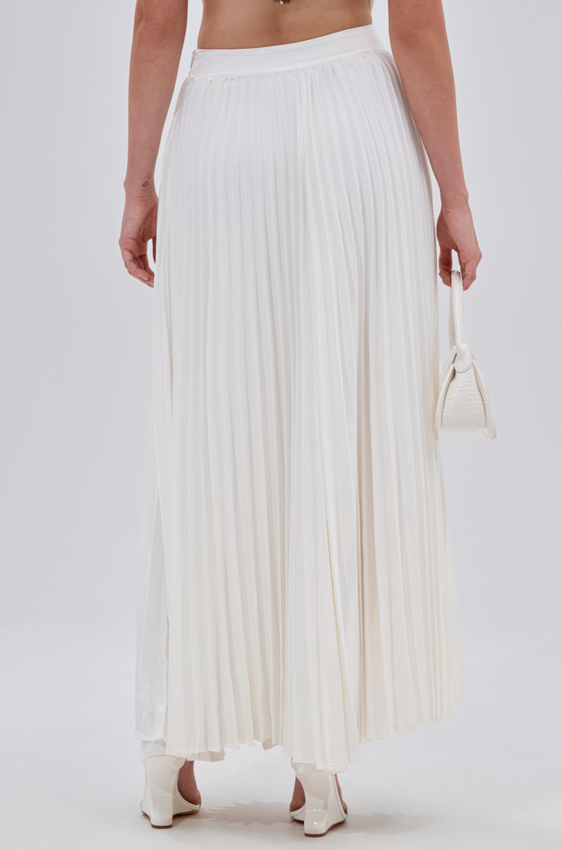 A person stands facing away in the SWEET THING PLEATED SATIN MAXI SKIRT styled with white heels and a small white handbag, against a plain light background. 3