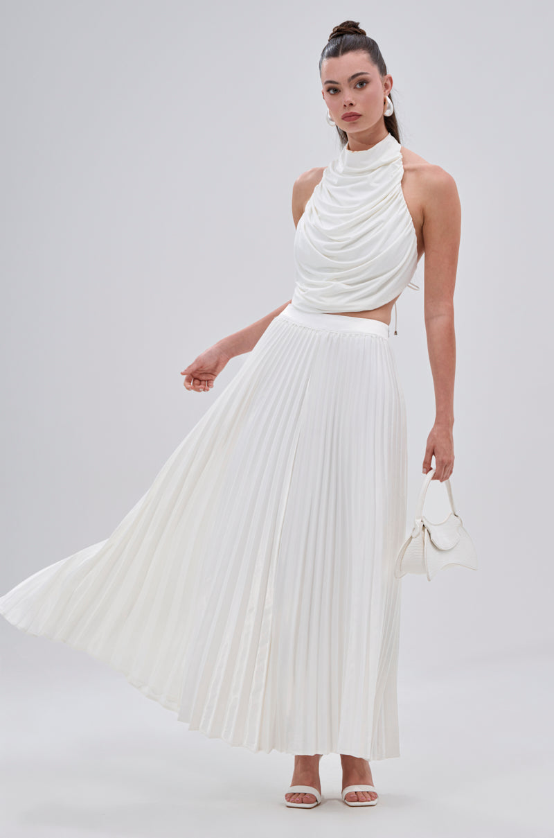 A woman poses against a plain background in the SWEET THING PLEATED SATIN MAXI SKIRT, styled with a draped halter top, small white handbag, and white sandals. 4