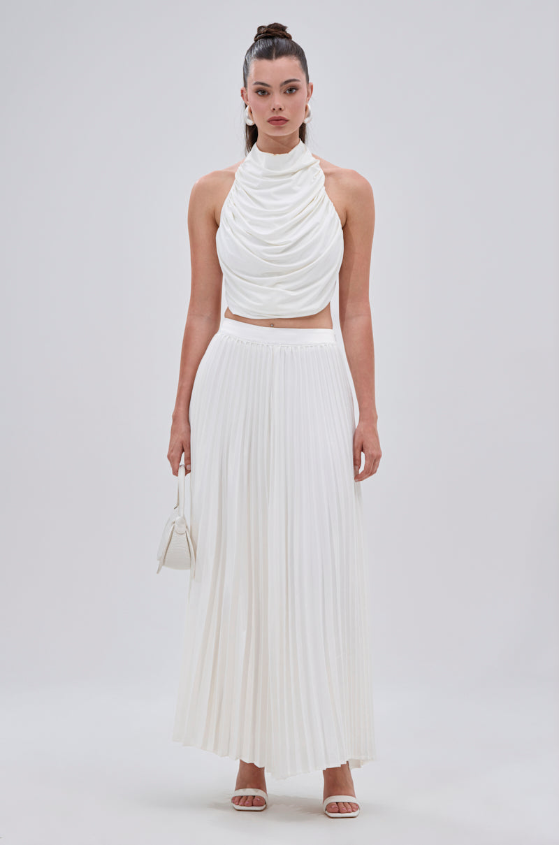 A woman stands against a plain background wearing a sleeveless white draped crop top and the SWEET THING PLEATED SATIN MAXI SKIRT. She accessorizes with a small white handbag, heeled sandals, and wears her hair in a high ponytail. 5
