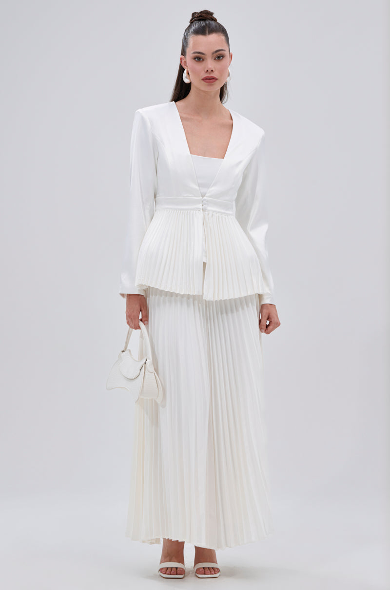 A woman wears the SWEET THING PLEATED SATIN MAXI SKIRT with a matching pleated jacket and white heels. She carries a small white handbag, her long hair styled in a sleek ponytail. 6