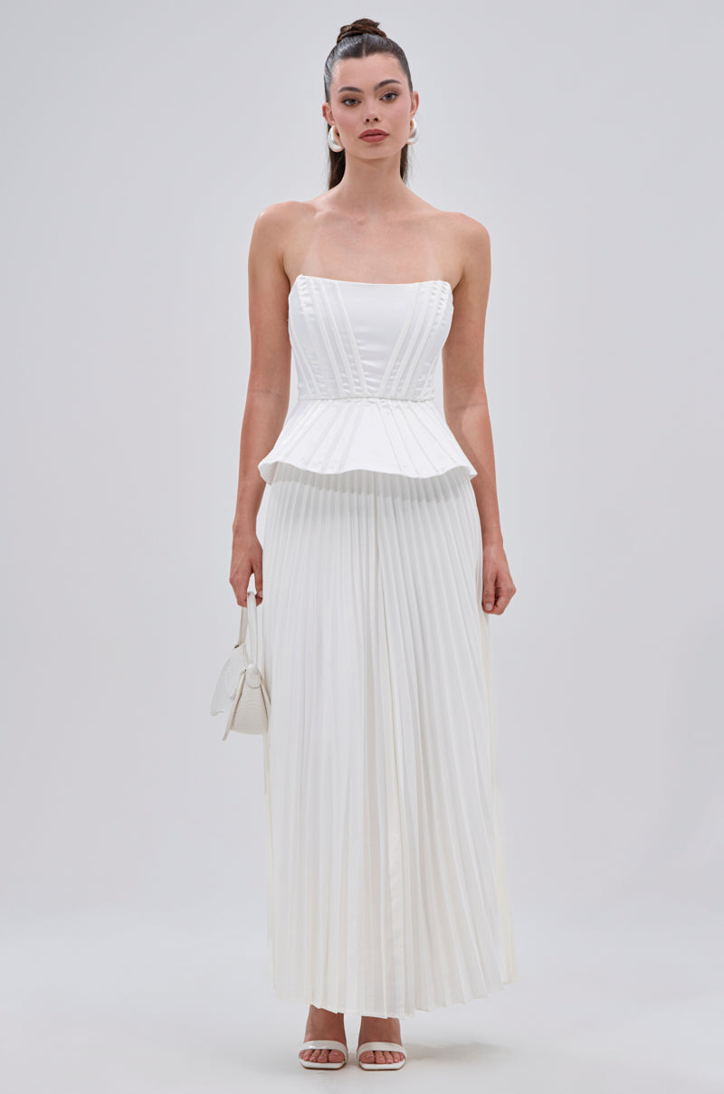 A woman wearing the SWEET THING PLEATED SATIN MAXI SKIRT with a strapless white dress, holding a small white handbag and wearing white heels, stands against a plain light background. 7