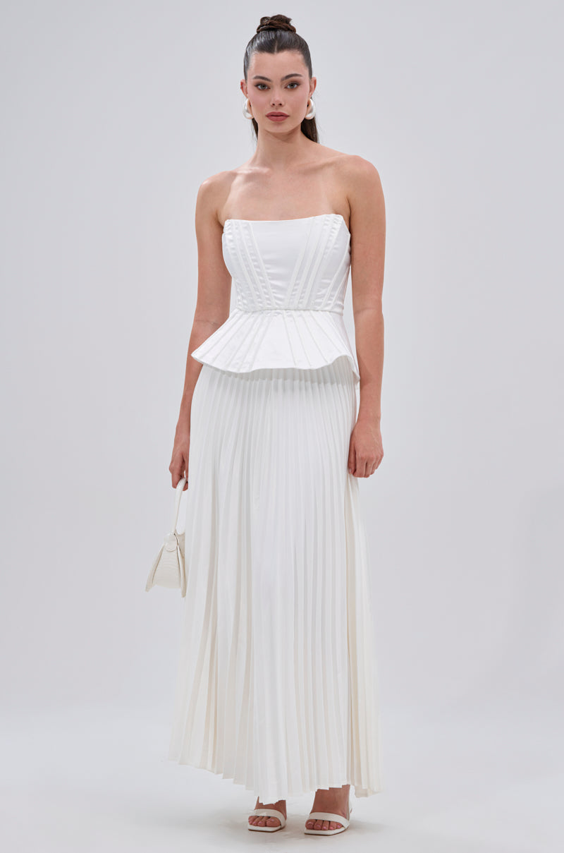 A woman stands against a plain background wearing the SWEET THING PLEATED SATIN MAXI SKIRT, styled with a strapless white corset top, white sandals, and a small white handbag. 8