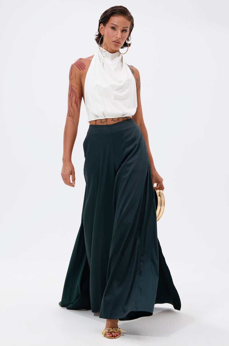 A person stands against a plain white background, wearing LOOK AT ME GO TROUSER WITH TAIL—satin-like, high-rise, wide-leg pants—paired with a sleeveless white halter top, open-toe heels, and holding a small gold handbag.