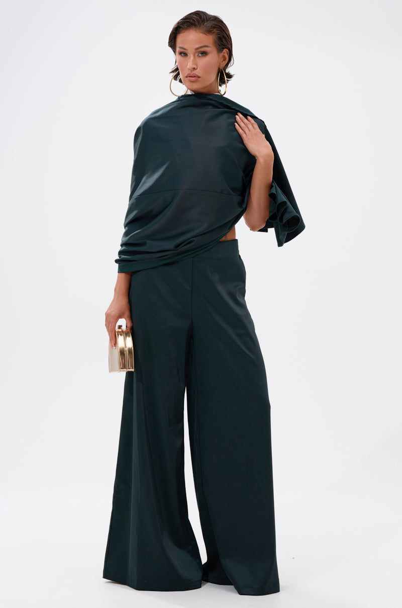 A woman poses against a white background in the LOOK AT ME GO TROUSER WITH TAIL, a high-rise wide-leg pant paired with a dark green asymmetrical satin top. She accessorizes with large hoop earrings and holds a gold clutch.