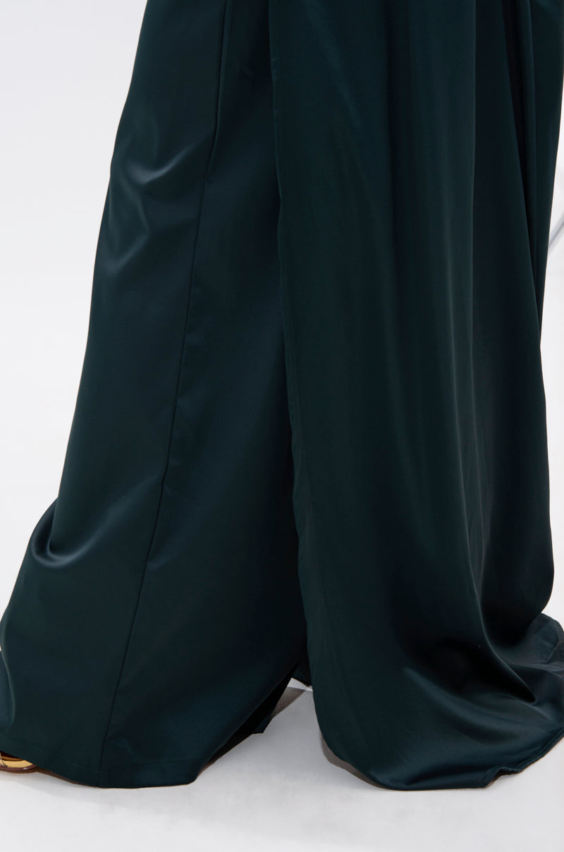 A close-up of the lower half of the LOOK AT ME GO TROUSER WITH TAIL in dark green satin-like fabric, draping elegantly to the floor against a plain, light background.