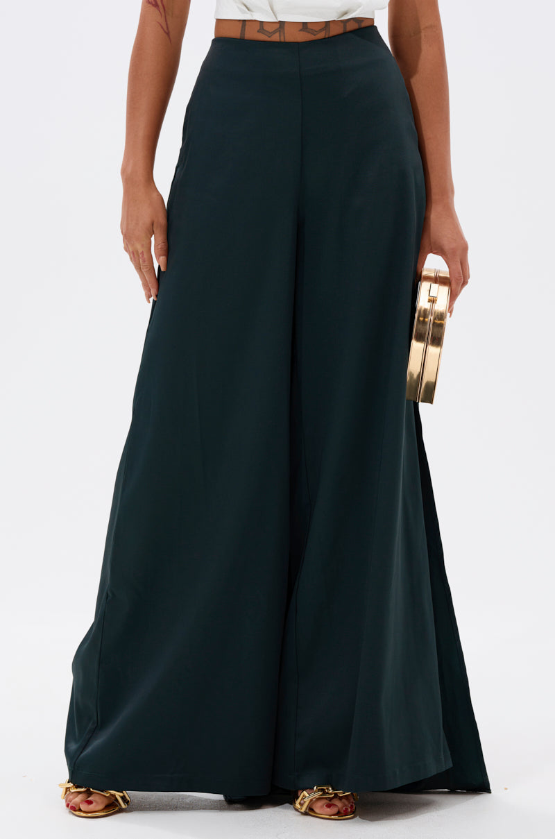 A person wearing the LOOK AT ME GO TROUSER WITH TAIL and open-toe sandals holds a round gold clutch, with their partially visible torso and arms at their sides against a plain white background.