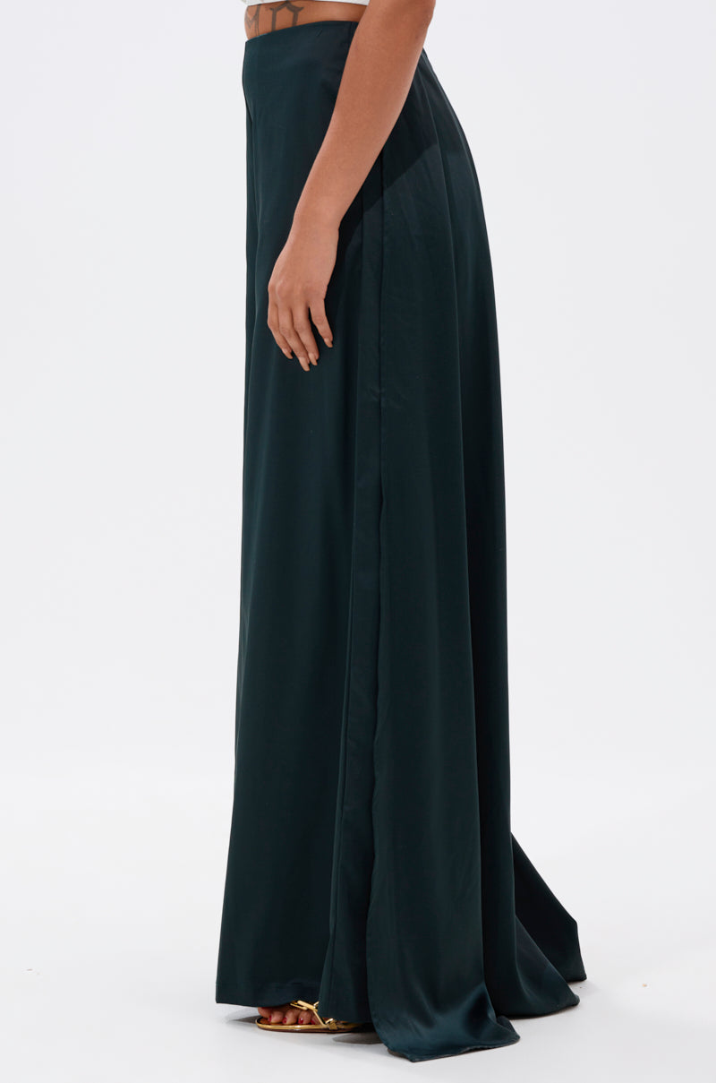 A person stands sideways against a plain white background wearing the LOOK AT ME GO TROUSER WITH TAIL, a long, dark green, high-waisted satin trouser featuring a subtle pleated maxi train, paired with open-toe heels.