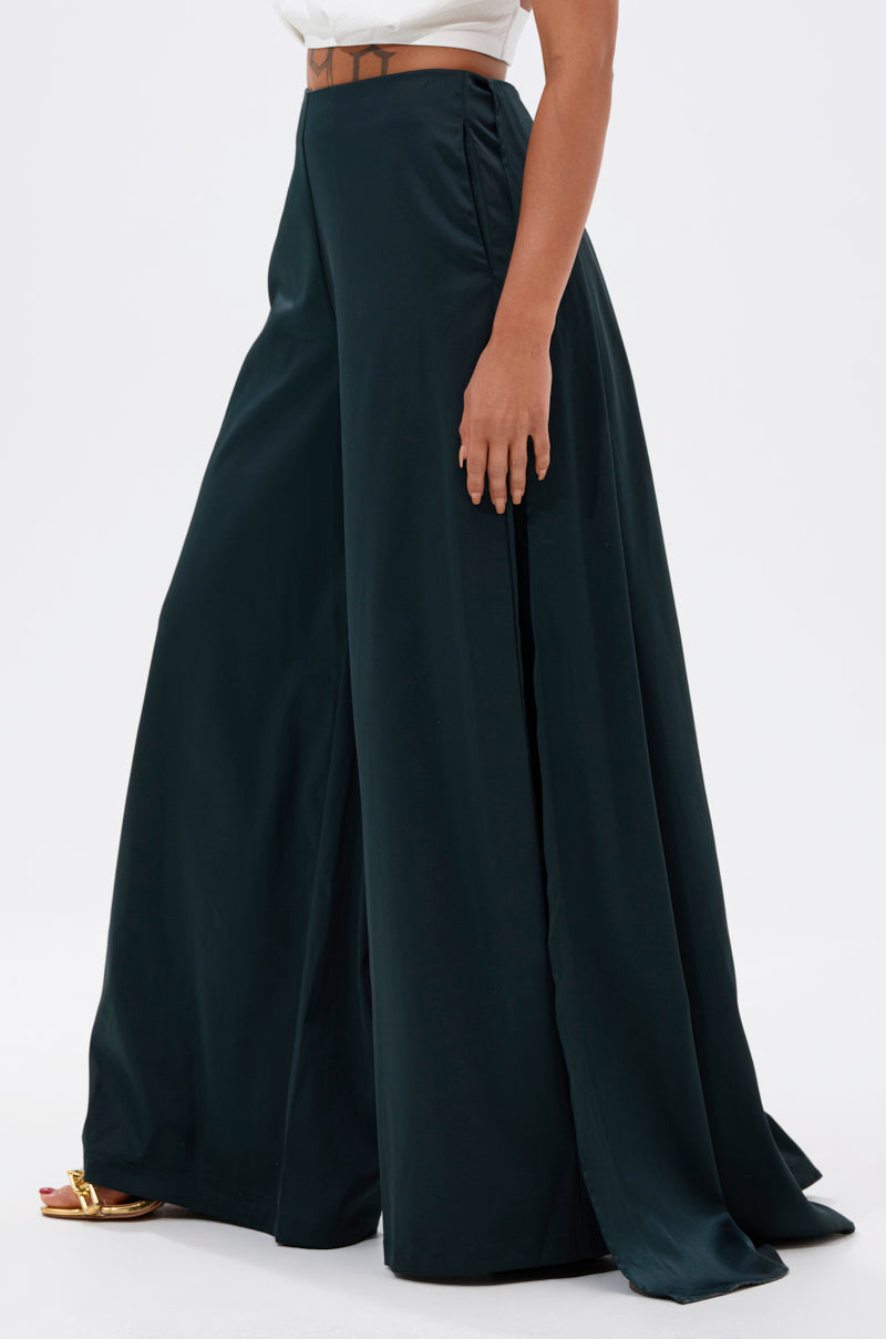 A person wearing the LOOK AT ME GO TROUSER WITH TAIL in dark green, styled with a white top and gold heeled sandals, stands against a plain white background.