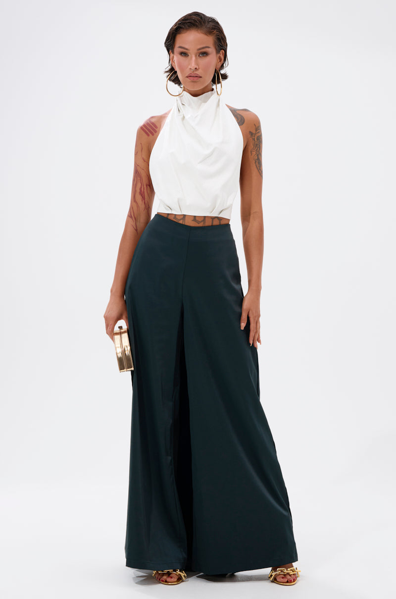 A woman with short hair wears a sleeveless white halter top and the LOOK AT ME GO TROUSER WITH TAIL, paired with heeled sandals, large hoop earrings, and a small gold clutch. Tattoos are visible on her arms.