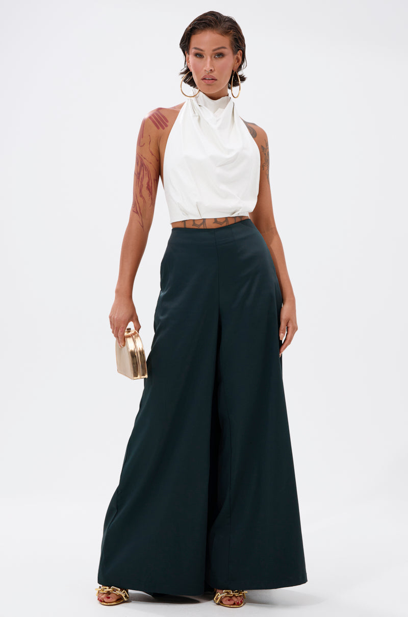 A woman poses confidently in the LOOK AT ME GO TROUSER WITH TAIL, paired with a sleeveless white halter top, gold sandals, hoop earrings, and a small gold clutch. Her styled hair and visible arm tattoos complete the bold look.
