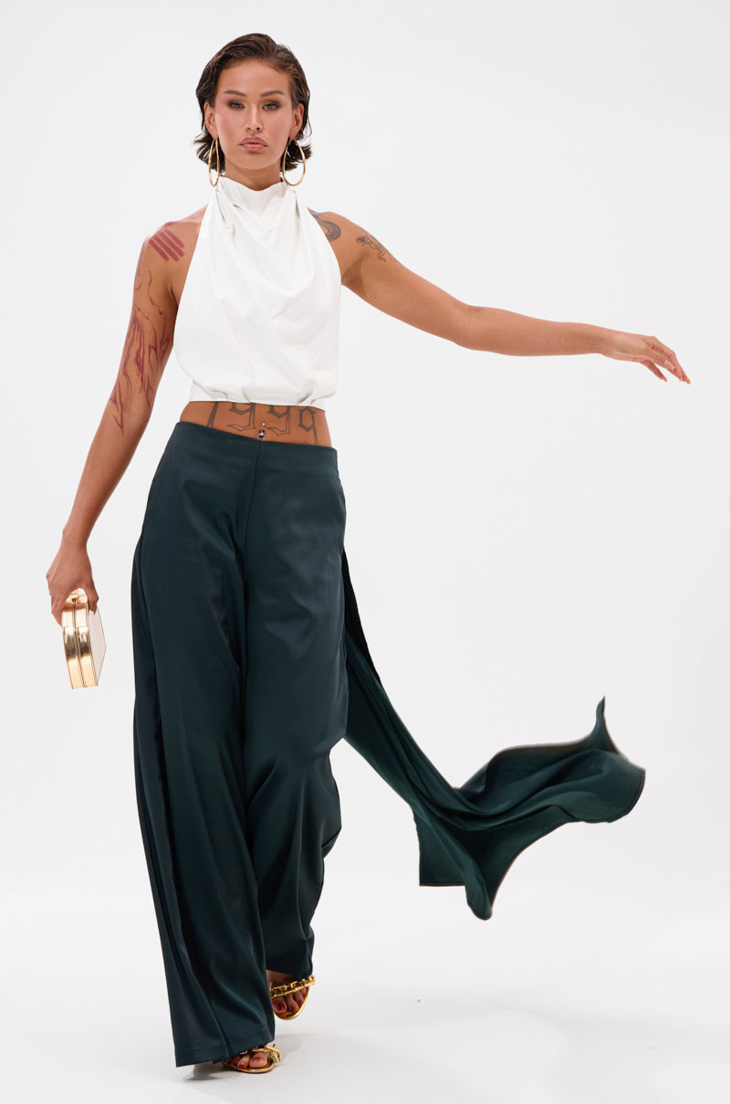 A person with short brown hair poses confidently in the LOOK AT ME GO TROUSER WITH TAIL and a sleeveless white top, holding a gold clutch. They have arm and torso tattoos, large hoop earrings, and sandals.