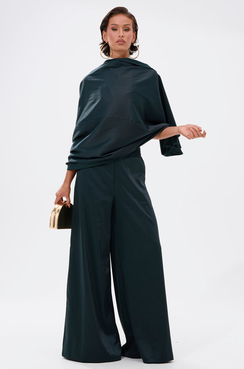 A woman poses confidently against a white backdrop, wearing the LOOK AT ME GO TROUSER WITH TAIL—a loose dark green satin-like set with wide sleeves and high-rise wide-leg trousers—paired with a small gold handbag and sleek, styled-back hair.