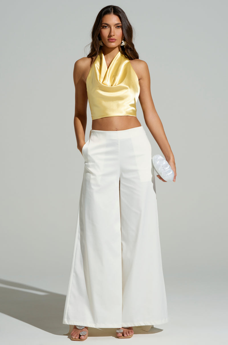 A woman stands confidently against a plain background, wearing a yellow satin halter neck top and LOOK AT ME GO WHITE TROUSER, featuring high-rise and wide-leg silhouette. She holds a small clutch in one hand while the other rests in her pocket, accessorized with strappy sandals.