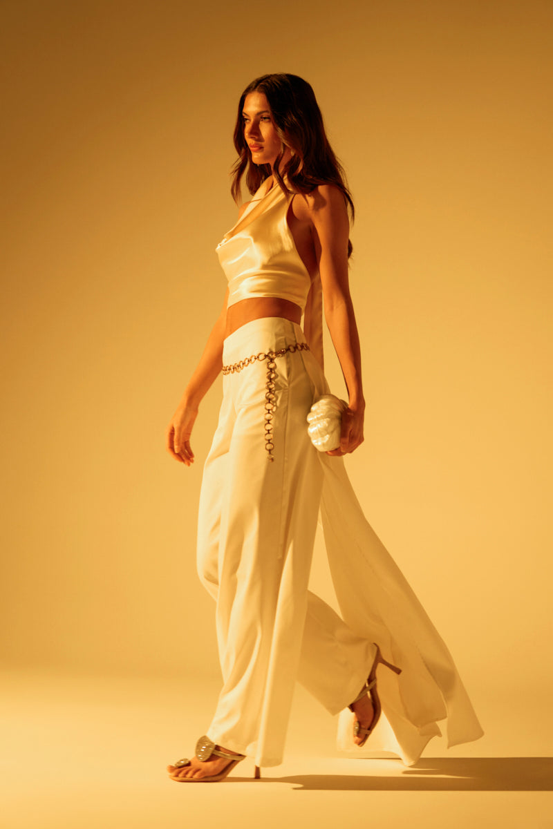 In warm studio lighting, a woman confidently walks wearing LOOK AT ME GO WHITE TROUSER, a sleek satin crop top, and wide-leg high-rise trousers. She's accessorized with a chain belt, carrying a small textured clutch and wearing heeled sandals. Her hair cascades in loose waves.