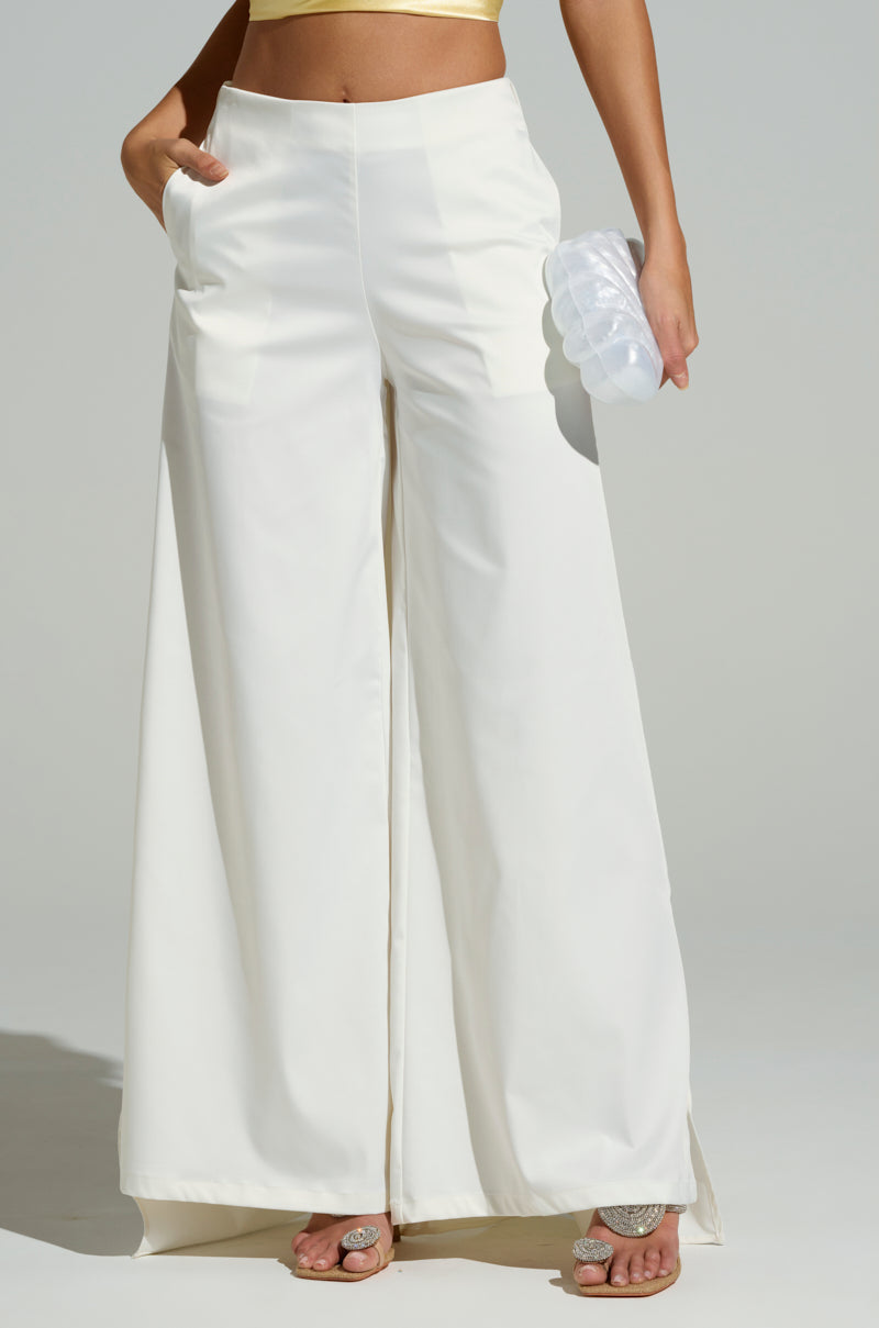 A person wearing the LOOK AT ME GO WHITE TROUSER with a wide-leg style and a light crop top is holding a translucent clutch, standing with one hand in their pocket. Their open-toed footwear features decorative stones against a plain gray background.