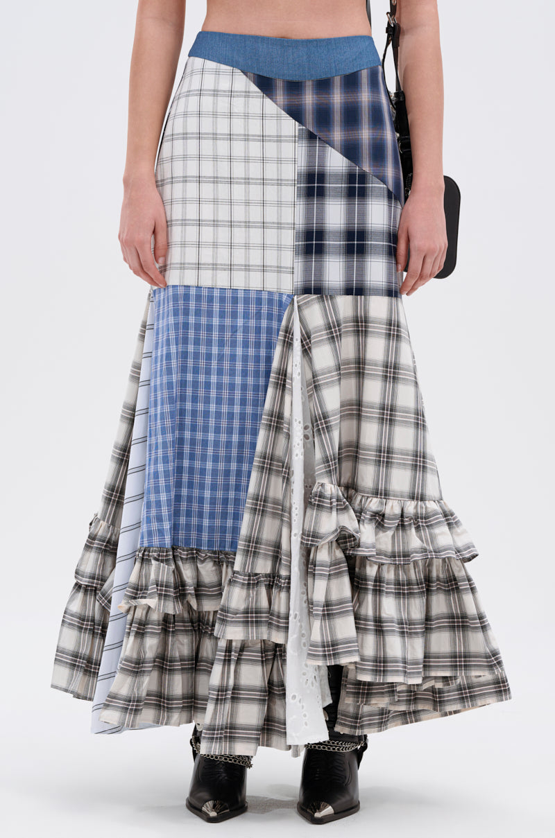 A person wears the OUT OF ALIGNMENT PLAID MIDI SKIRT, featuring blue, white, and black plaid with ruffled hem details and a denim high-rise waistband, styled with black boots.