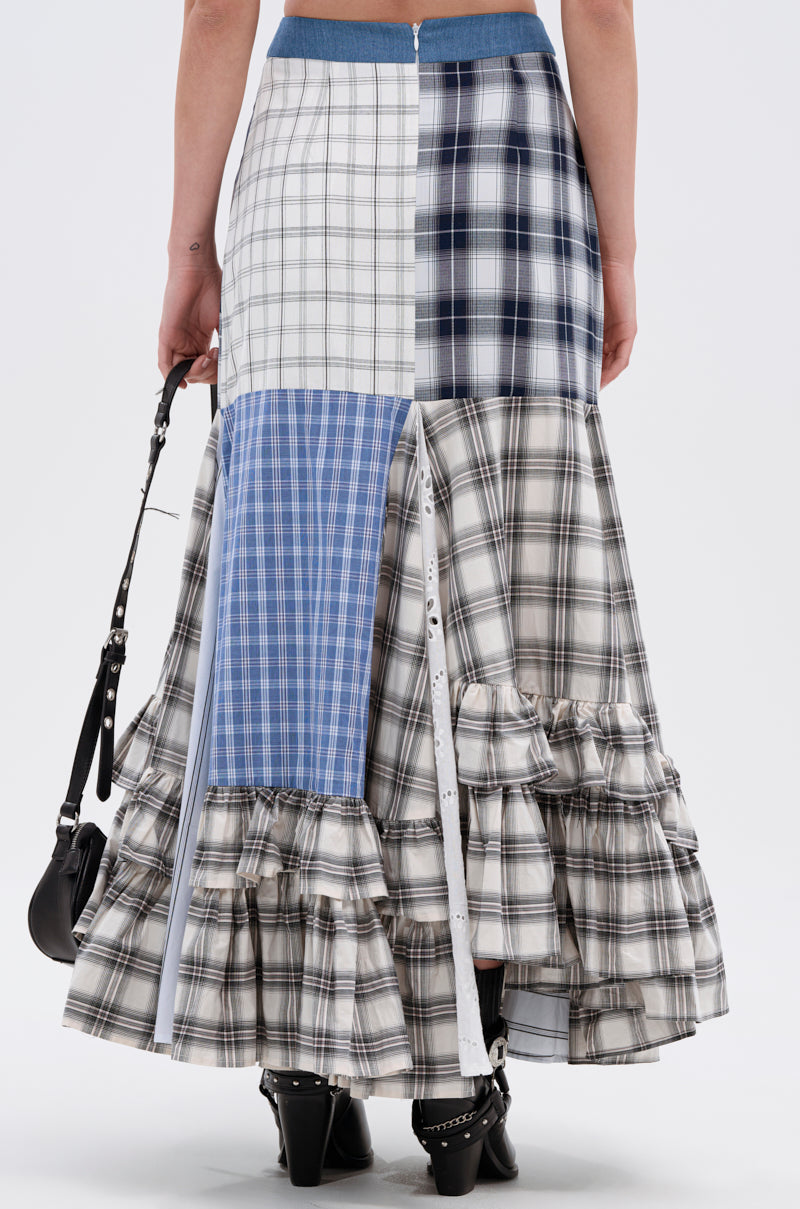 A woman is seen from the back wearing the OUT OF ALIGNMENT PLAID MIDI SKIRT, a patchwork plaid midi with ruffled layers in blue, white, and black tones and a denim high-rise waistband, holding a small black handbag.