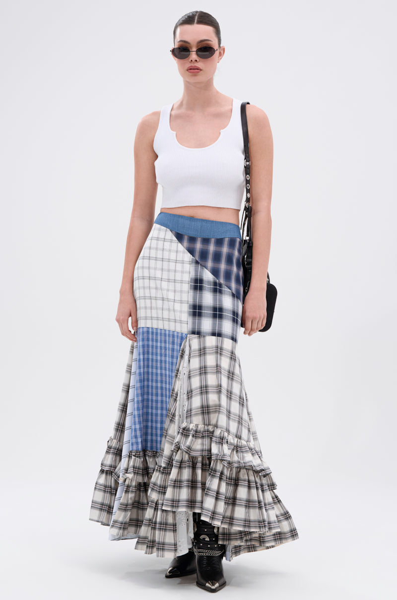 A woman stands against a plain background wearing sunglasses, a white cropped tank top, black ankle boots, a black shoulder bag, and the OUT OF ALIGNMENT PLAID MIDI SKIRT with ruffles and a denim high-rise waistband.