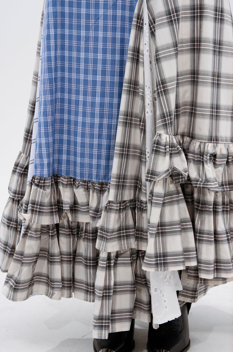 Close-up of two ruffled skirts: the OUT OF ALIGNMENT PLAID MIDI SKIRT in blue and white, and another in black, white, and gray; both styled with black boots. A touch of white eyelet fabric shows between the skirts.