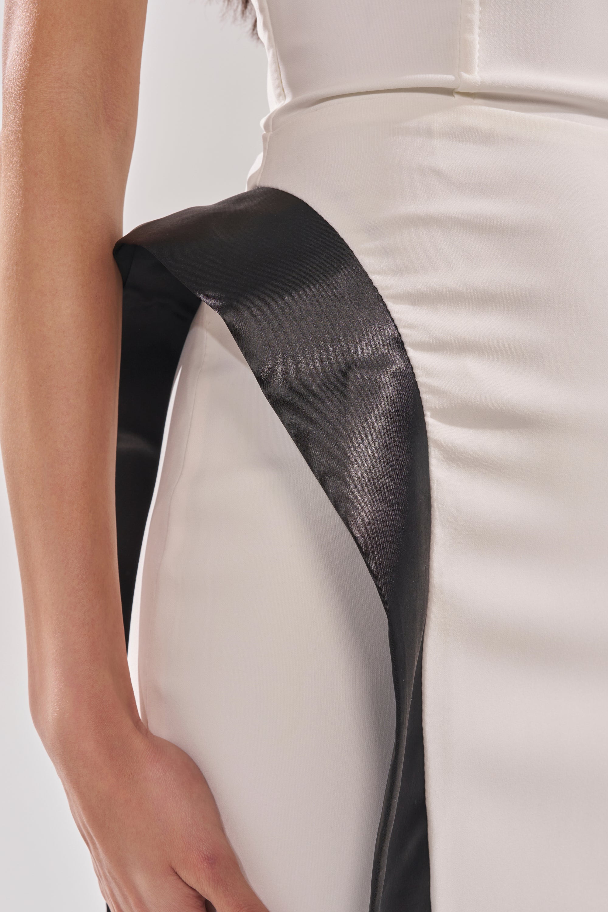 A close-up of a person wearing the CHIC AND SOPHISTICATED STRUCTURED MIDI DRESS IN WHITE, a strapless bodycon style with stretch fabric and a shiny black sash draped over the hip, their hand resting by their side. 9