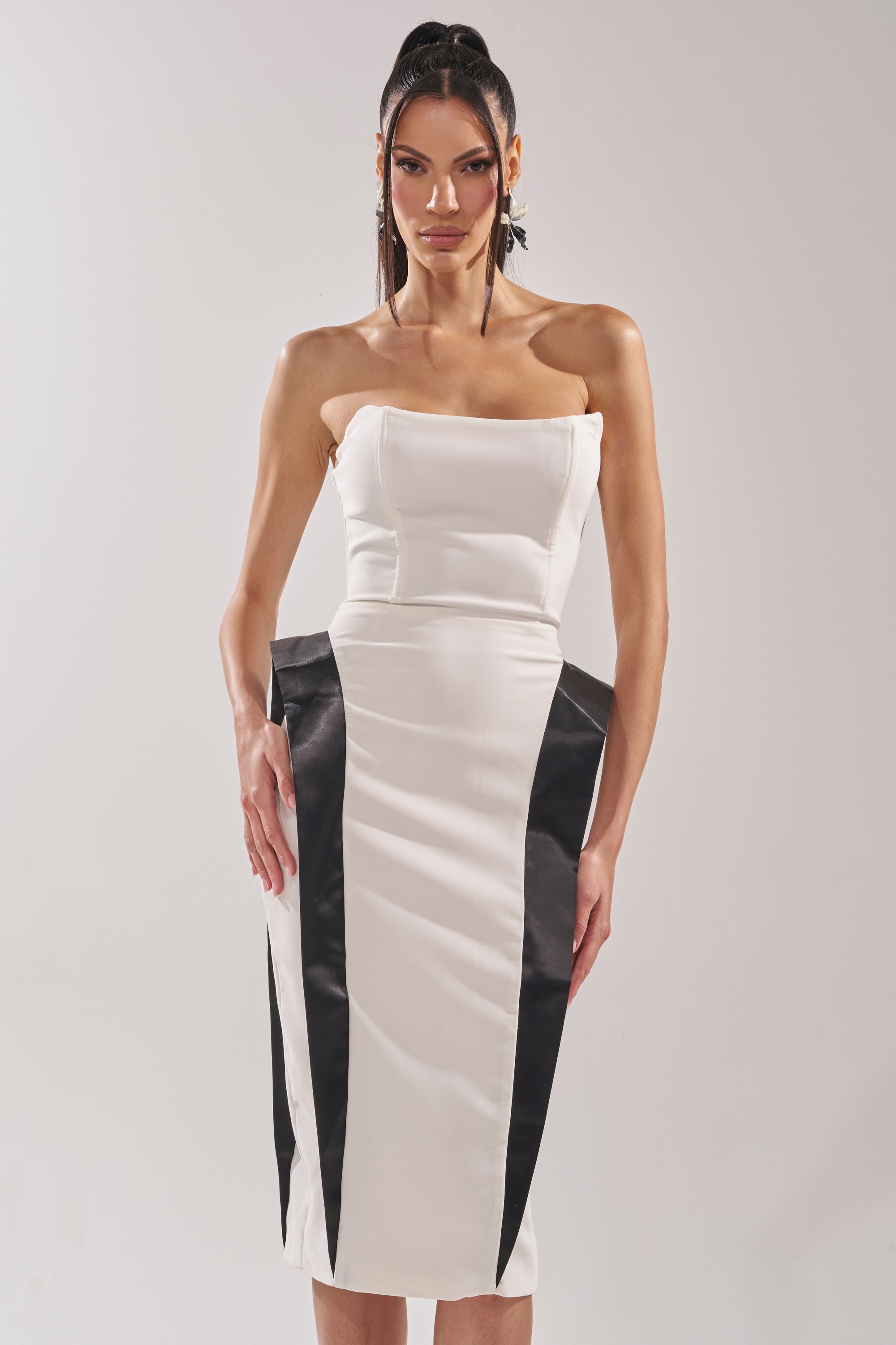 A woman wears the CHIC AND SOPHISTICATED STRUCTURED MIDI DRESS IN WHITE, featuring strapless styling and black side panels for an hourglass effect. She stands against a plain light background, facing the camera with arms by her sides. 1