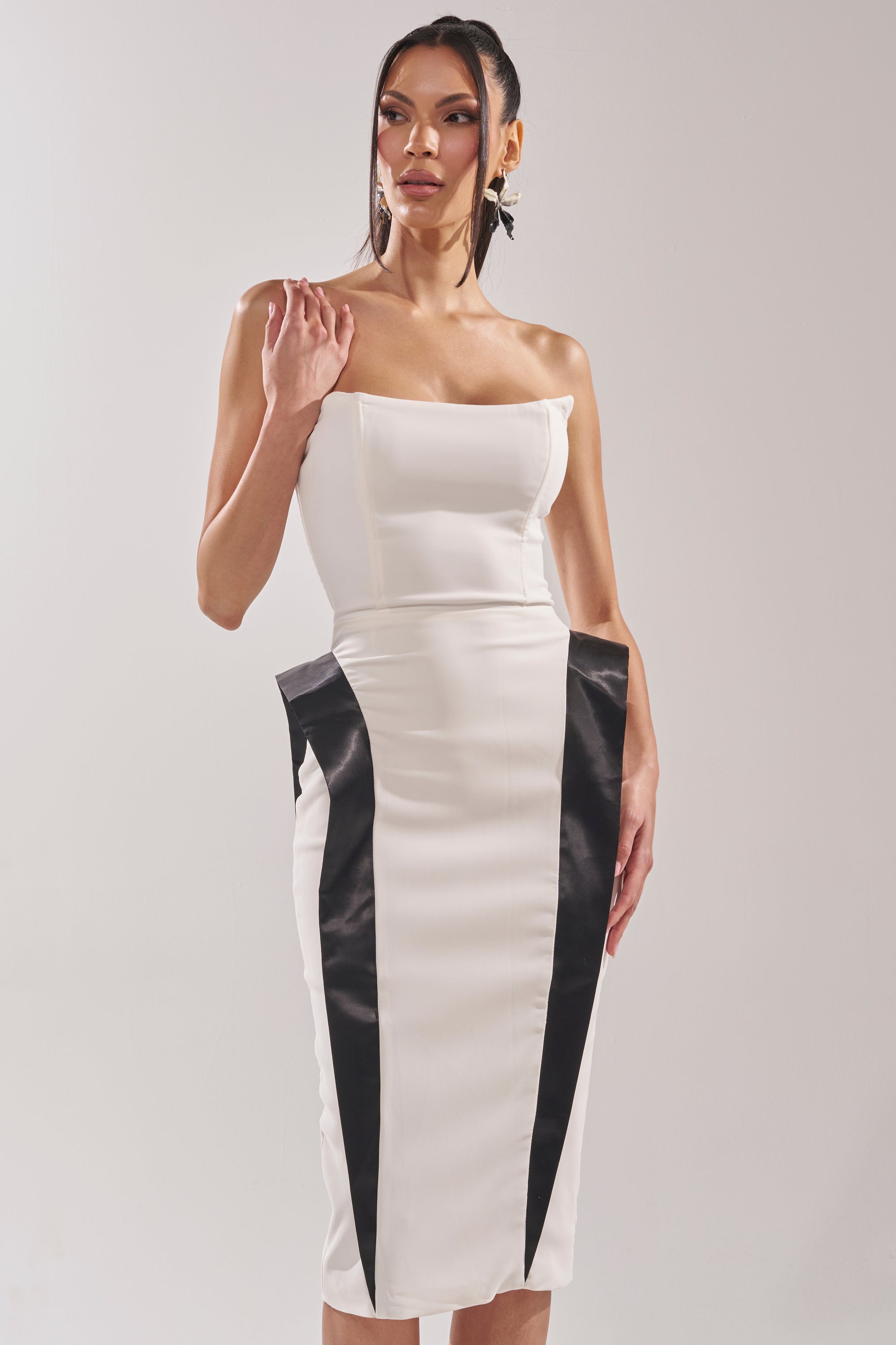 A woman models the CHIC AND SOPHISTICATED STRUCTURED MIDI DRESS IN WHITE, featuring bold black side panels that create a striking hourglass illusion. She poses against a light background with her hair in a sleek ponytail. 2