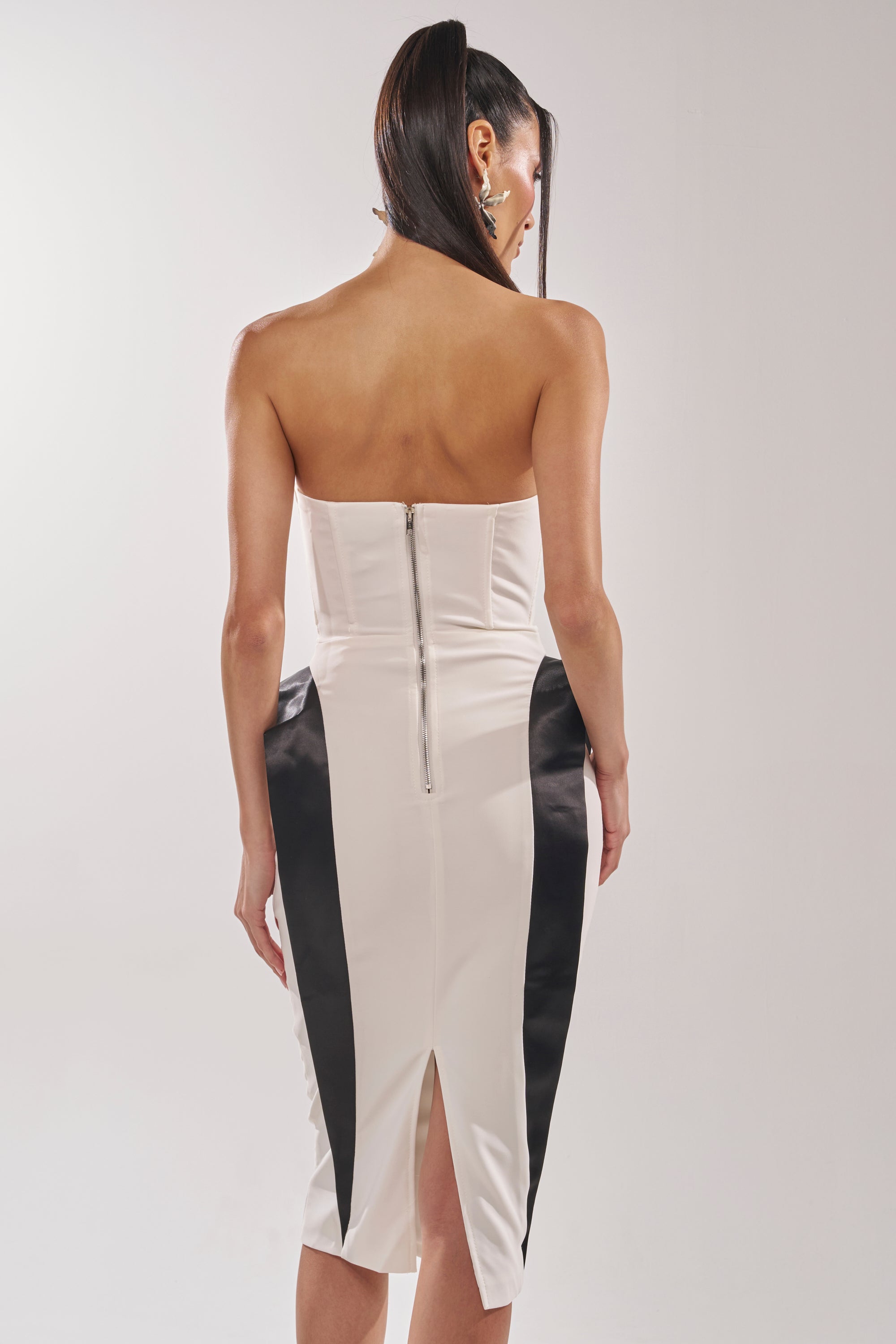A woman with straight, long hair models the CHIC AND SOPHISTICATED STRUCTURED MIDI DRESS IN WHITE, featuring black contrast panels and a back zipper for an hourglass effect as she poses with her back to the camera in a bright studio. 4
