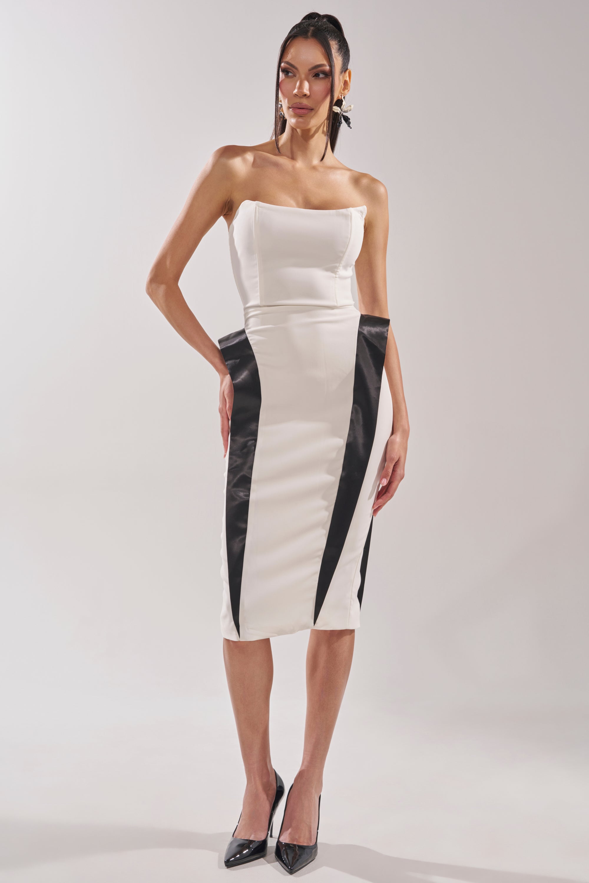 A woman models the CHIC AND SOPHISTICATED STRUCTURED MIDI DRESS IN WHITE with black contrast panels, creating an hourglass silhouette, paired with black pointed-toe heels against a plain light background. 5