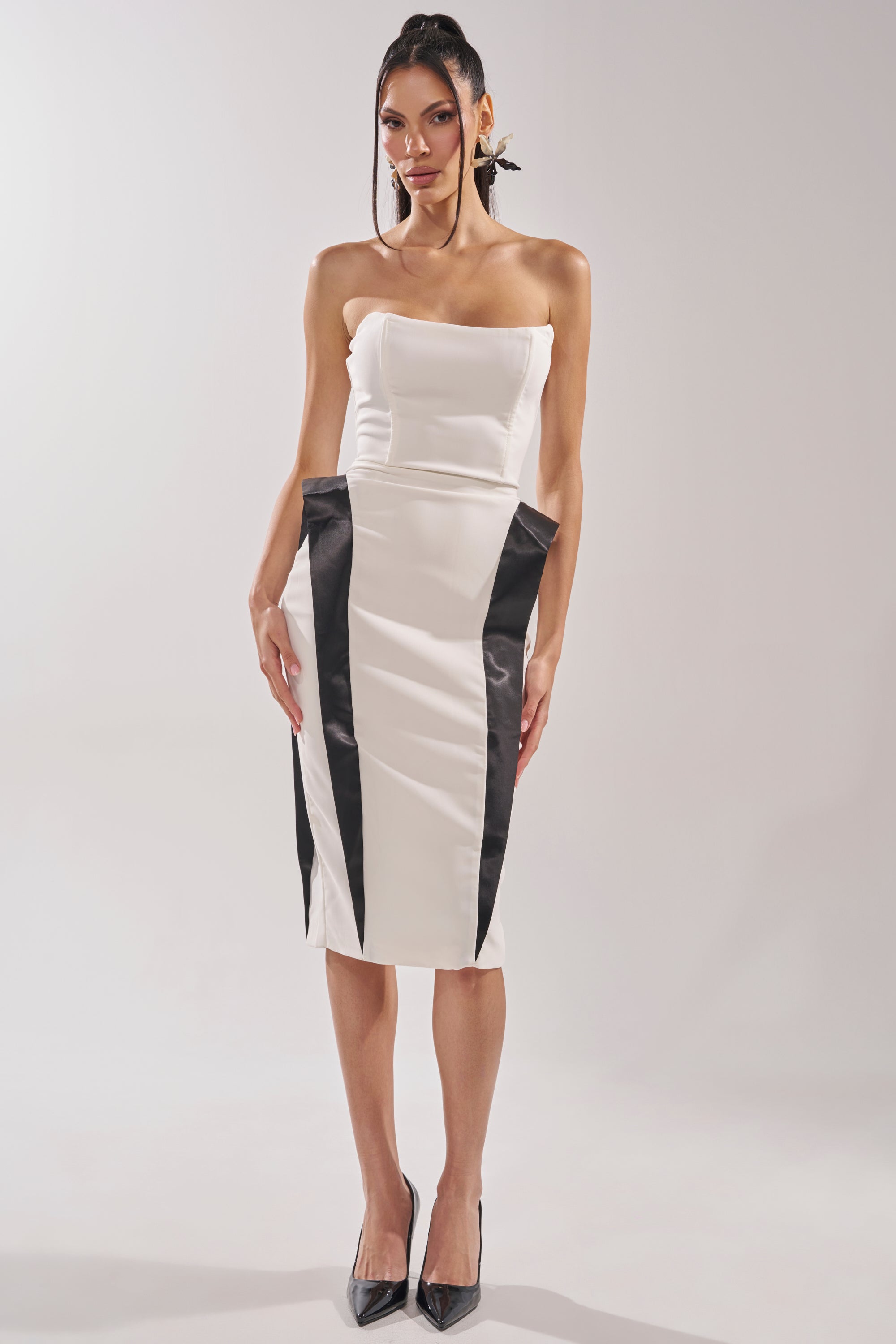 A woman wears the CHIC AND SOPHISTICATED STRUCTURED MIDI DRESS IN WHITE, a strapless bodycon midi with black vertical panels creating an hourglass effect, paired with black pointed-toe heels against a plain light background. 6