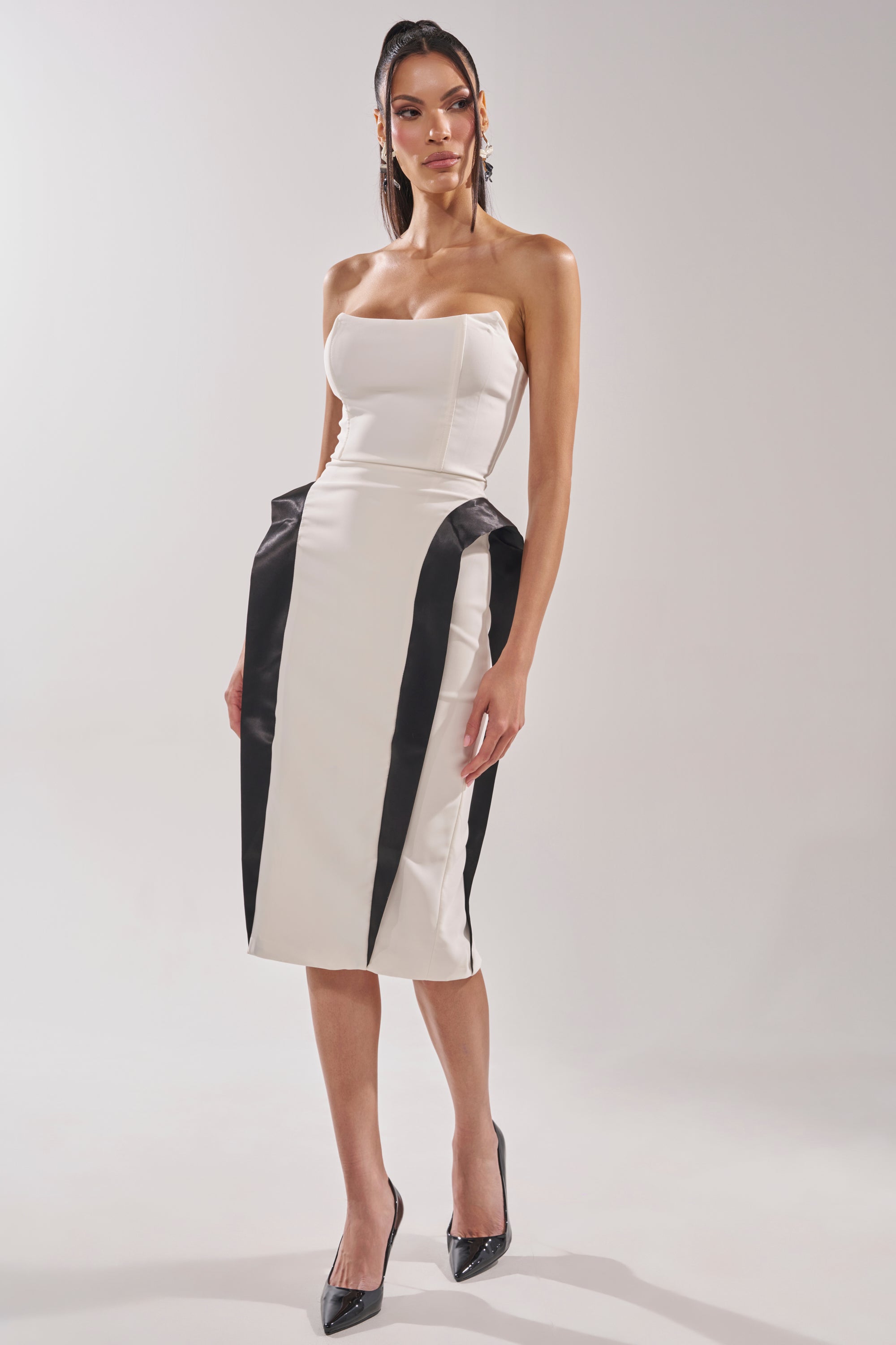 A woman models the CHIC AND SOPHISTICATED STRUCTURED MIDI DRESS IN WHITE, featuring a strapless bodycon fit with bold black vertical accents and an hourglass effect, styled with black pointed heels against a plain light background. 7