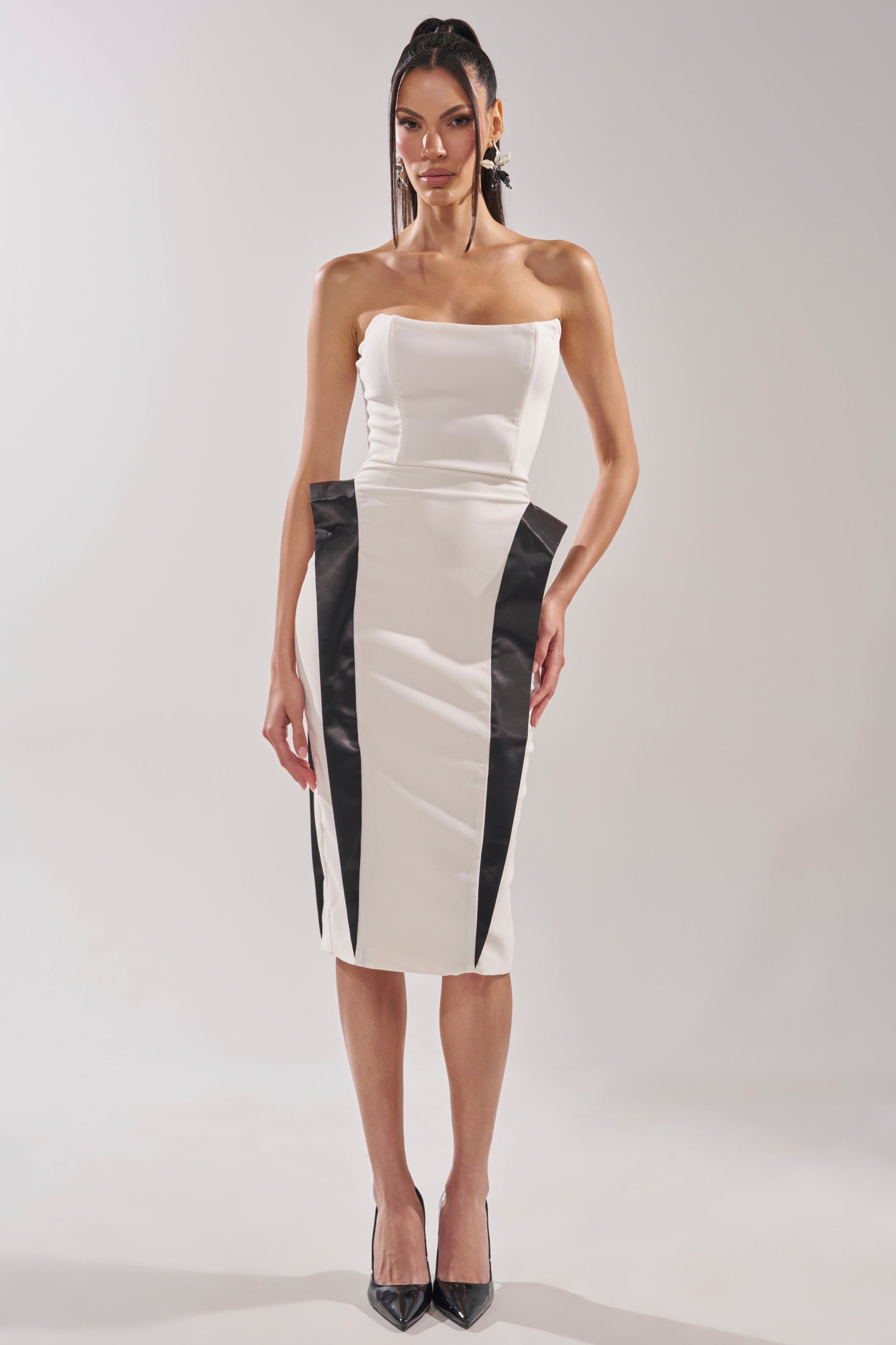 A woman wears the CHIC AND SOPHISTICATED STRUCTURED MIDI DRESS IN WHITE, a strapless bodycon style with black vertical panels for an hourglass effect, paired with straight hair and black heels, against a plain light background. 8