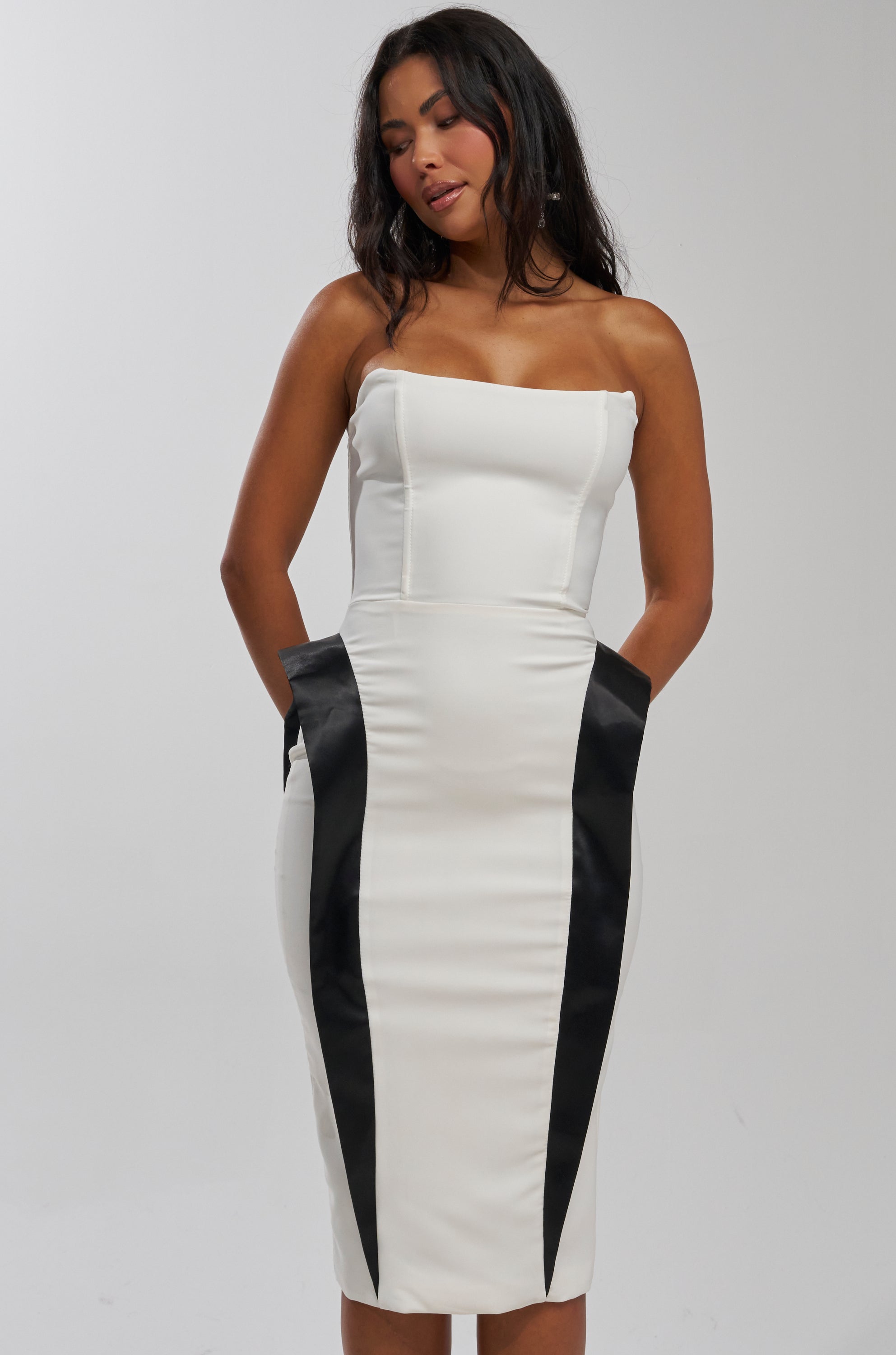 A woman stands against a plain background wearing the CHIC AND SOPHISTICATED STRUCTURED MIDI DRESS IN WHITE, its black contrast side panels and strapless bodycon fit creating an effortless hourglass effect as she looks down with a relaxed expression.
