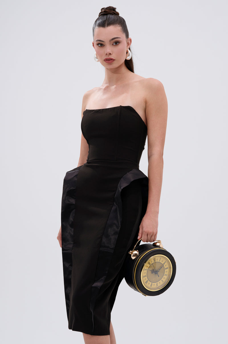 A woman wears the CHIC AND SOPHISTICATED STRUCTURED MIDI DRESS, a strapless black midi with hip accents, paired with a vintage clock-inspired round handbag and large earrings. Her hair is in a high bun against a plain light gray background.