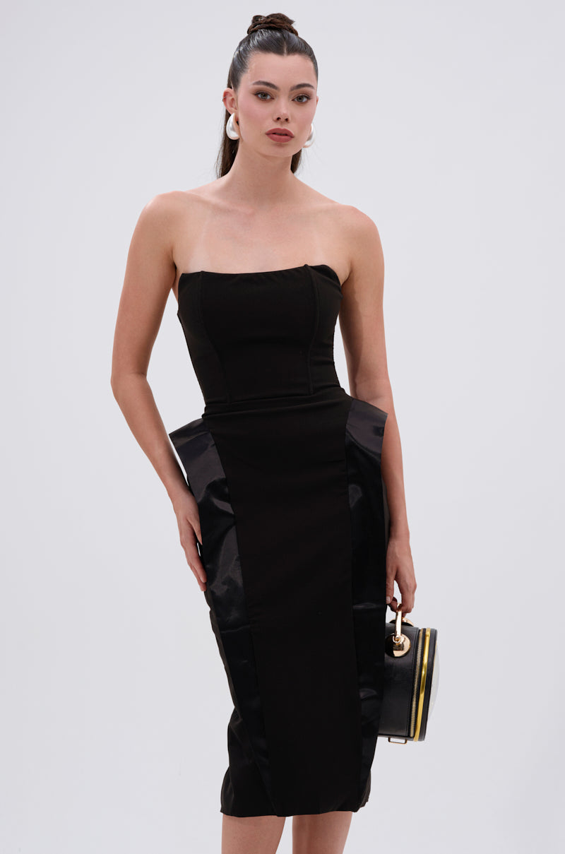 A woman wears the CHIC AND SOPHISTICATED STRUCTURED MIDI DRESS, featuring a tailored silhouette and structured hip accents, holding a small black handbag and confidently facing the camera against a light background.