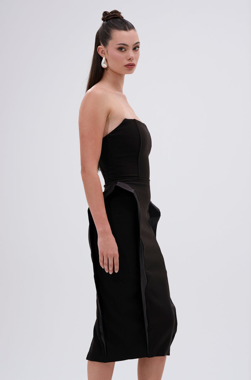 A woman with long dark hair styled half-up wears the CHIC AND SOPHISTICATED STRUCTURED MIDI DRESS—strapless, black, with structured hip accents—and statement earrings, standing against a plain light gray background.