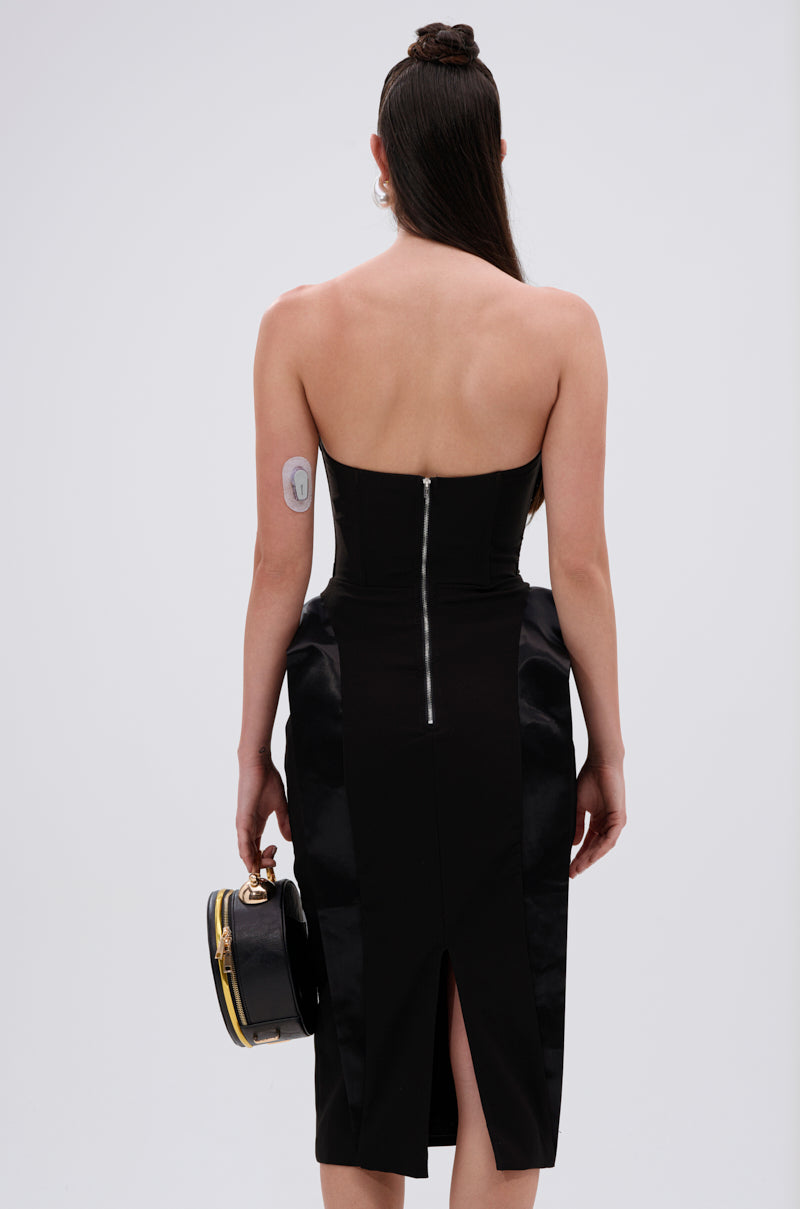 A person with long dark hair stands facing away, wearing the CHIC AND SOPHISTICATED STRUCTURED MIDI DRESS in black, featuring a tailored fit, back zipper, and slit. They hold a round black handbag and have a white medical patch on their upper arm.