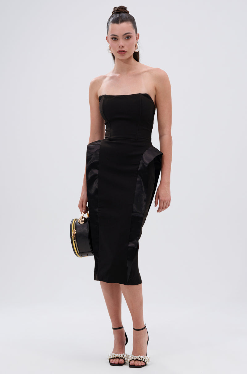 A woman poses against a plain background in the CHIC AND SOPHISTICATED STRUCTURED MIDI DRESS, featuring a tailored silhouette and structured hips. She accessorizes with a round black handbag, pearl-embellished heels, and a high ponytail.