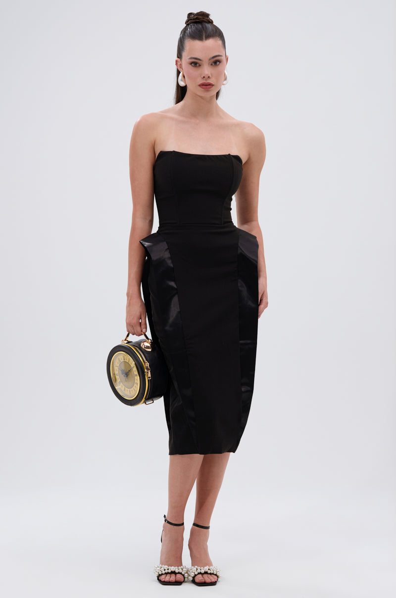 A woman stands against a plain background wearing the CHIC AND SOPHISTICATED STRUCTURED MIDI DRESS, styled with a round clock purse, strappy pearl-detailed heels, and her hair in a high ponytail.