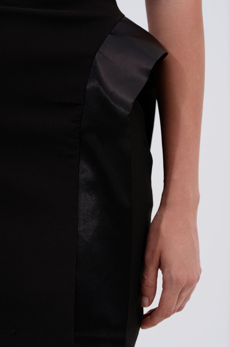 A close-up of a person wearing the CHIC AND SOPHISTICATED STRUCTURED MIDI DRESS, featuring a black finish with shiny satin panel and structured hip accents, their arm and hand resting by their side.
