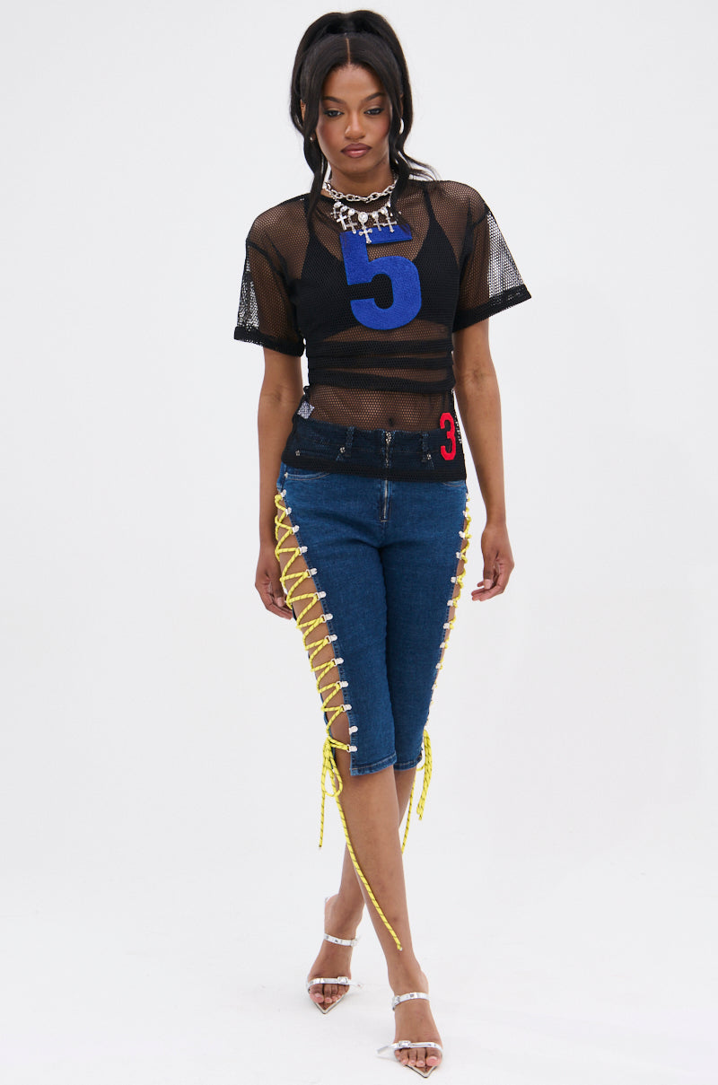 A woman wears the CUTE AS CAN BE LACE-UP DENIM CAPRI with yellow lace-up sides, paired with a black mesh top featuring a large blue number 5, clear strap heels, and a chunky silver necklace against a plain white background. 0