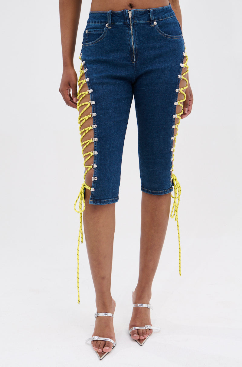Someone wears the CUTE AS CAN BE LACE-UP DENIM CAPRI—mid-rise blue denim capri shorts with yellow lace-up sides and silver grommets—paired with clear high-heeled sandals. 1