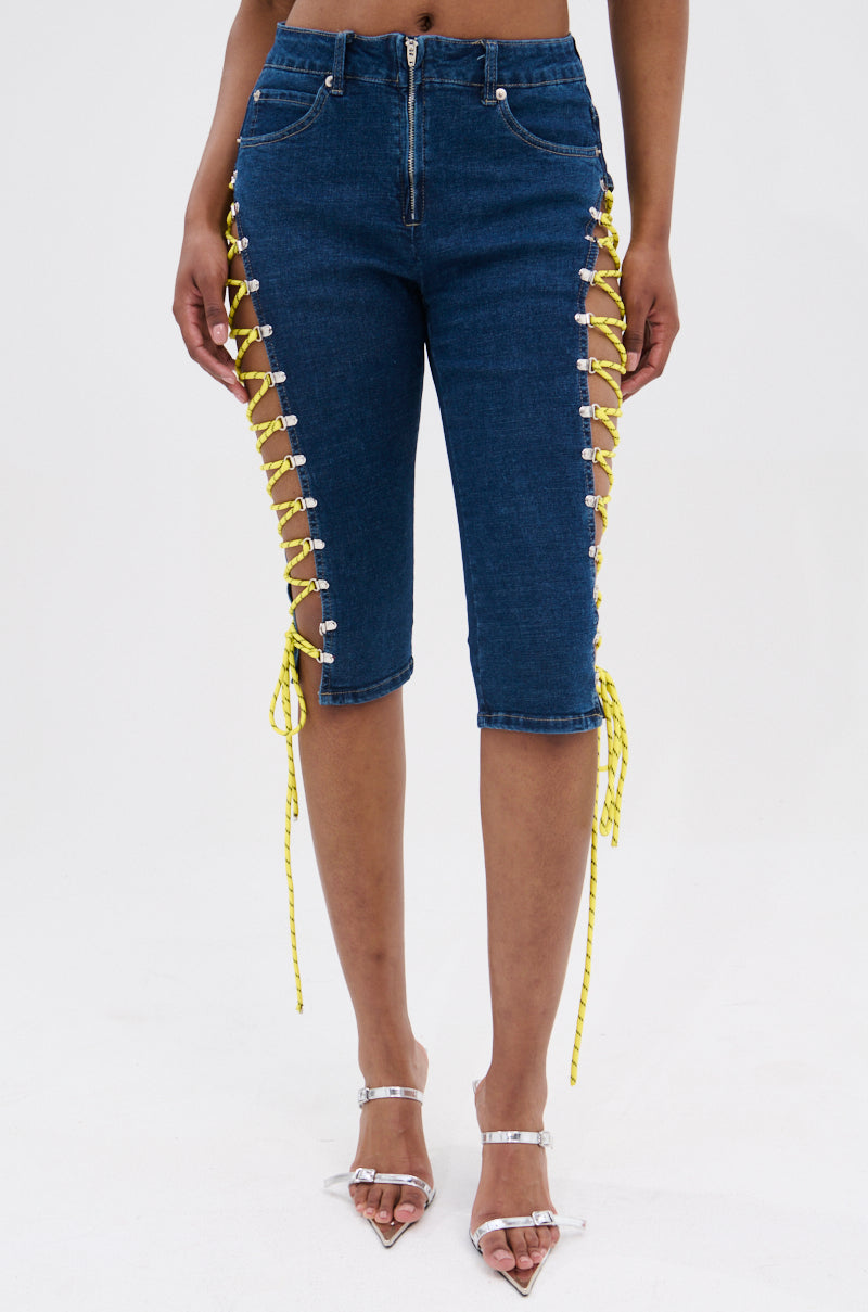 A person wearing the CUTE AS CAN BE LACE-UP DENIM CAPRI—mid-rise blue denim capri shorts with yellow lace-up sides—paired with silver high-heeled sandals, stands against a plain white background. 2