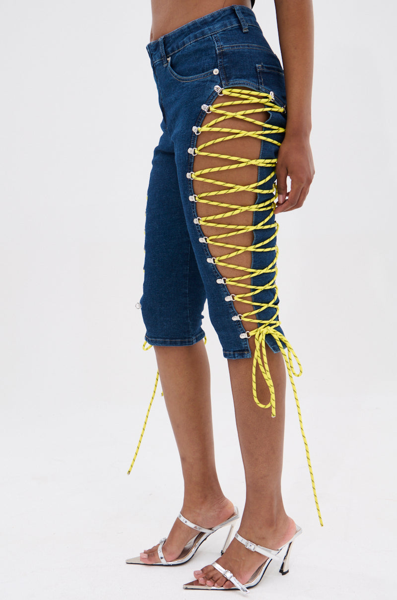 A model wears the CUTE AS CAN BE LACE-UP DENIM CAPRI—mid-rise blue denim capris with open side seams and bright yellow lace-up details—styled with white heeled sandals, set against a plain background. 3