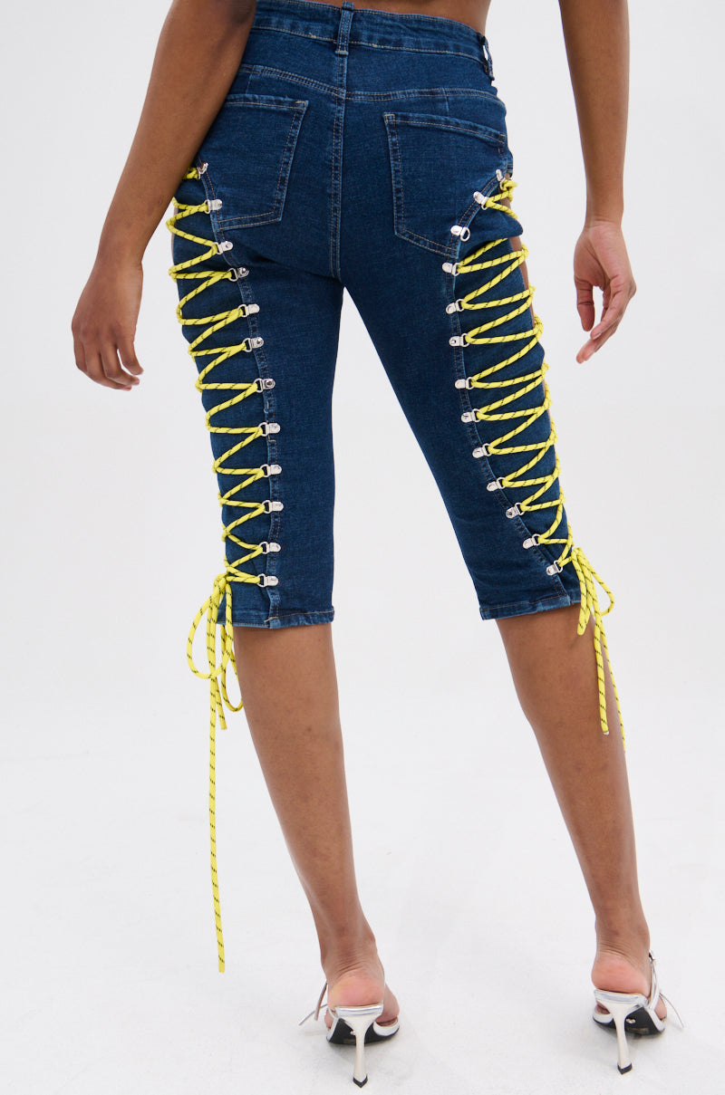 A person models the CUTE AS CAN BE LACE-UP DENIM CAPRI—mid-rise blue denim capris with yellow lace-up details on the back—paired with white heeled sandals, standing against a plain white background. 4