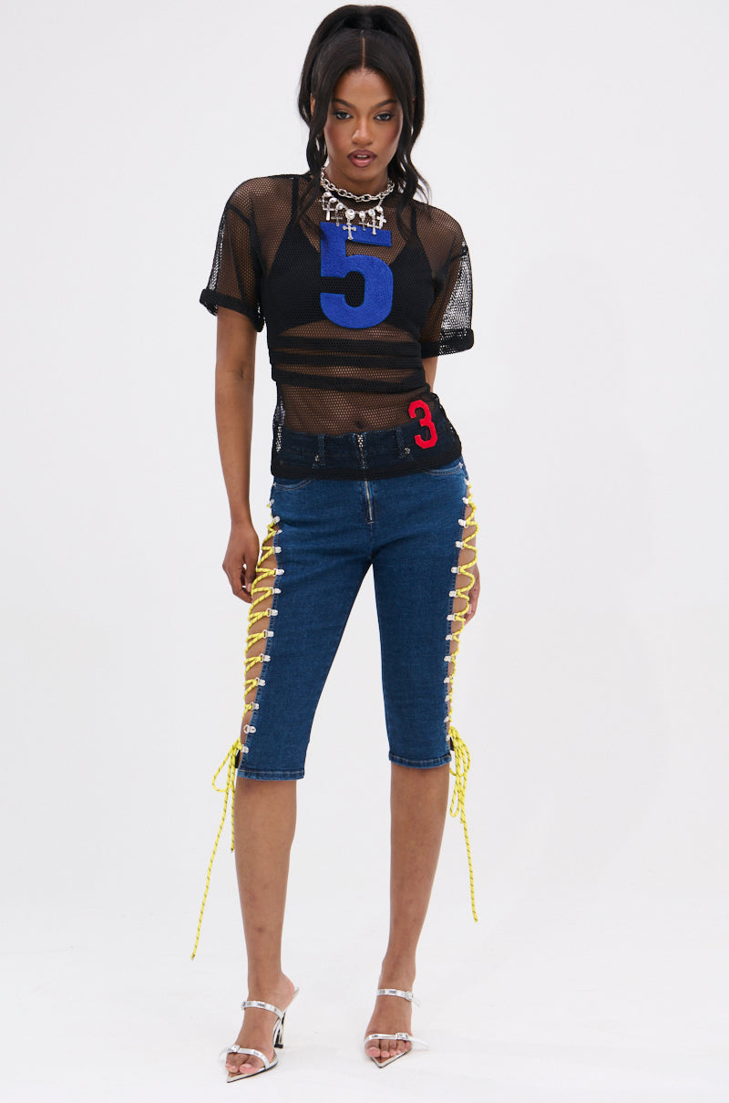 A woman wears the CUTE AS CAN BE LACE-UP DENIM CAPRI, a black mesh top with a bold blue 5, clear heels, and a chain necklace. Her hair is styled in loose waves as she poses against a white background. 5