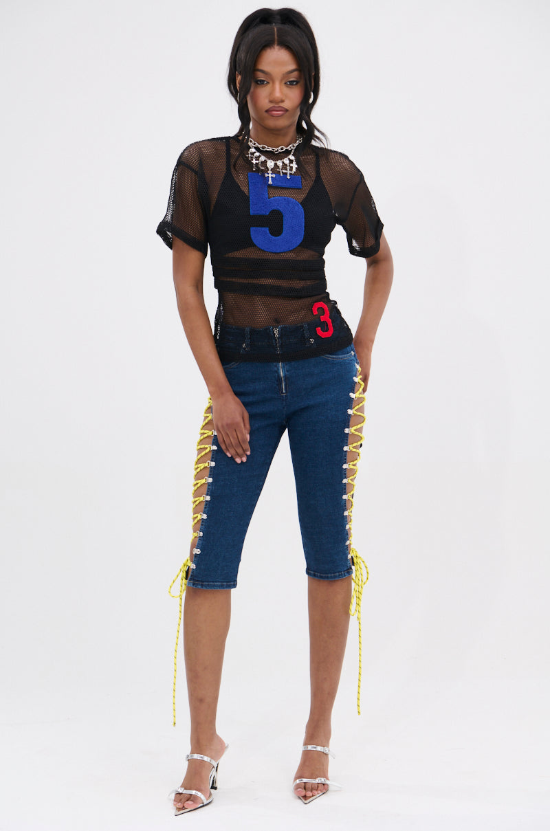 A woman stands against a white background wearing the CUTE AS CAN BE LACE-UP DENIM CAPRI, a black mesh top with blue and red numbers, silver heels, and a chunky necklace. 6