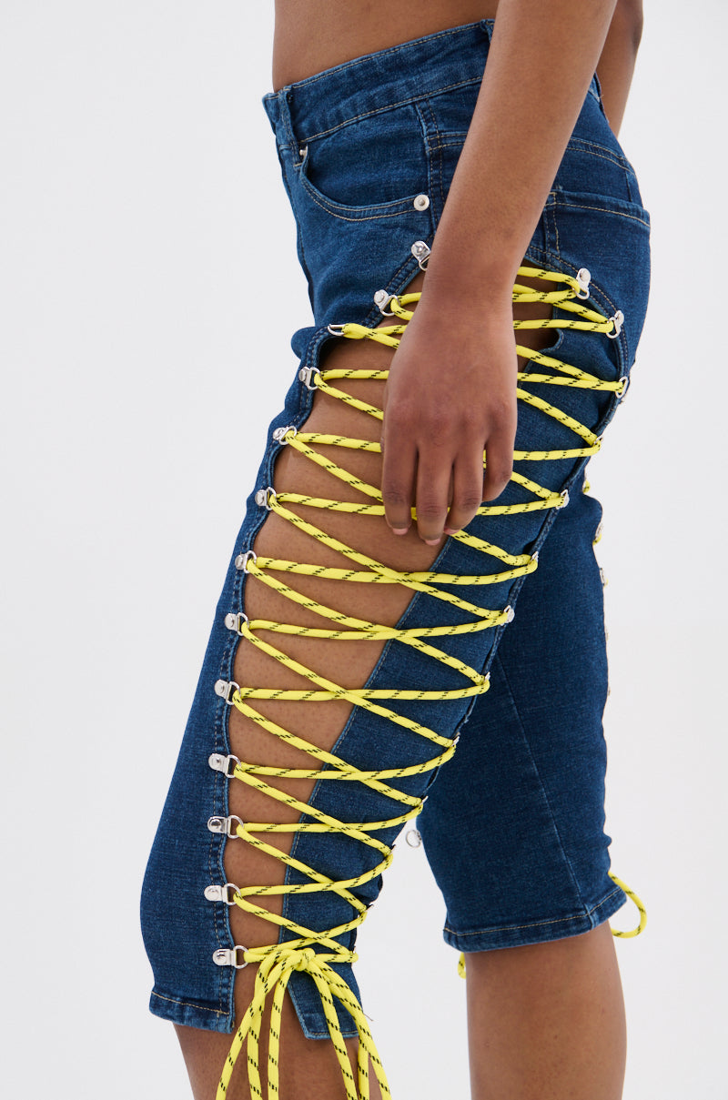 A person wears the CUTE AS CAN BE LACE-UP DENIM CAPRI, a mid-rise capri featuring open sides and bright yellow crisscross lace-up details that reveal skin. 7