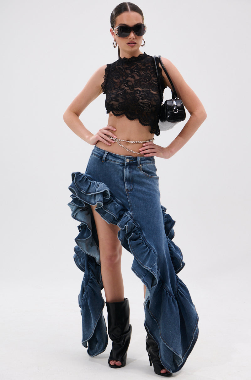 A woman poses in a black lace crop top, the DONT MESS WITH MY HEART DENIM RUFFLE SKIRT with dramatic edges and a front slit, black heeled boots, sunglasses, hoop earrings, a chain belt, and carries a small black handbag.